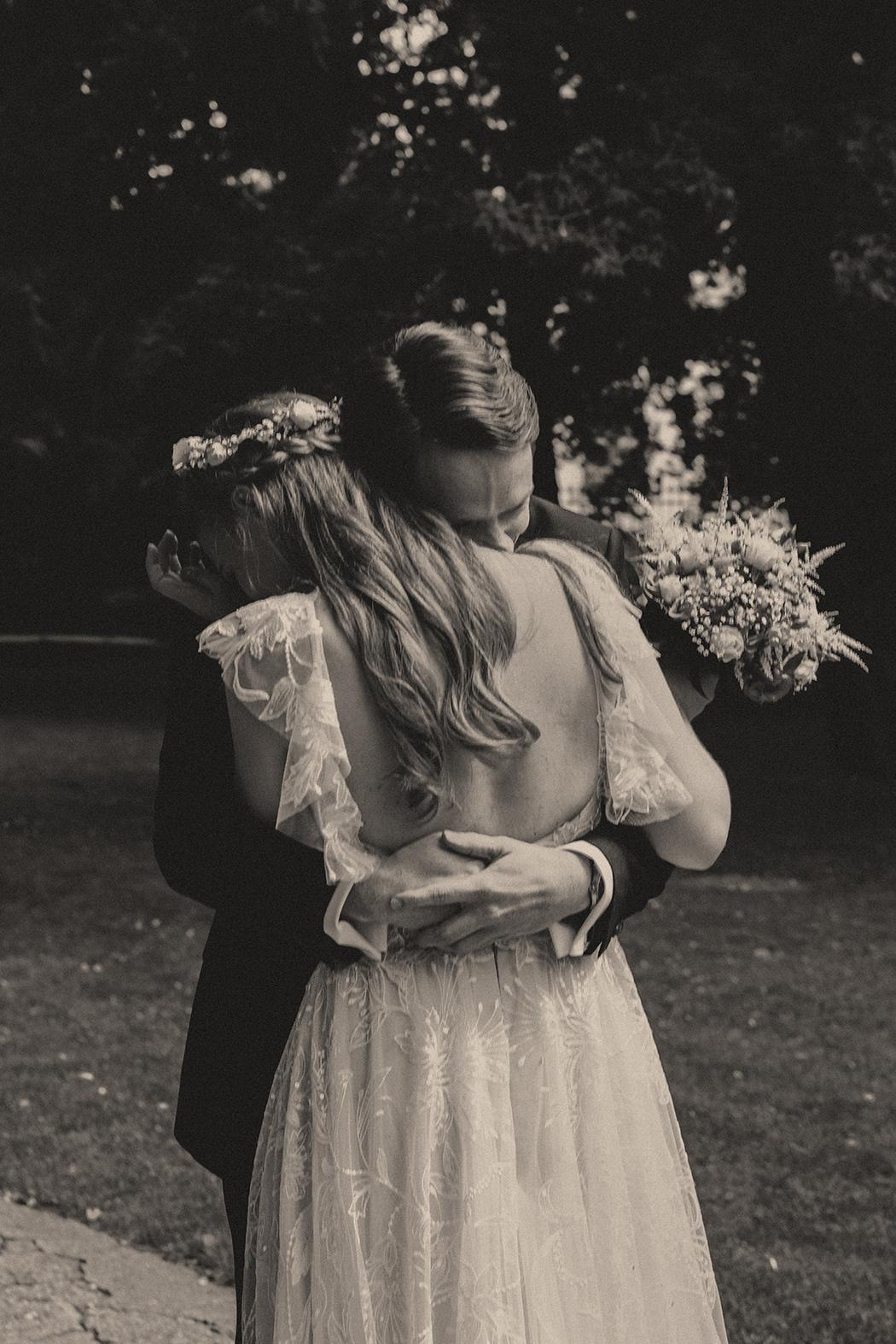 Das Schwarz-Weiß-Foto zeigt ein Brautpaar in inniger Umarmung. Die Braut trägt ein langes, zartes Brautkleid und eine Blumenkrone. Der Bräutigam umarmt sie von hinten, sein Kopf ruht auf ihrer Schulter. Ein Brautstrauß liegt neben ihnen auf dem Boden. Der Hintergrund ist unscharf und zeigt eine dunkle, bewaldete Umgebung. Die Szene wirkt emotional und intim.