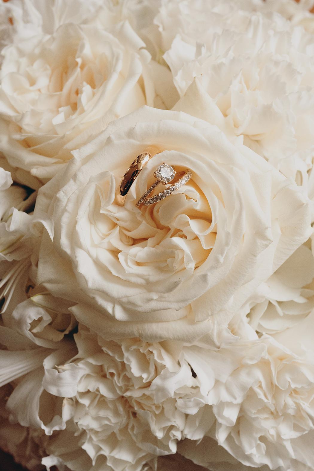 The image shows two gold wedding rings nestled amidst a bouquet of cream-colored roses and carnations. The rings are placed closely together, with one featuring a larger diamond in the center. The flowers form a soft, romantic backdrop that gently highlights the rings. The overall image is elegant and understated, with a focus on details and textures.