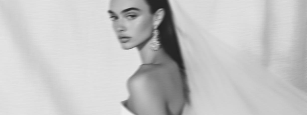 Black and white close-up portrait of a bride. The woman is positioned in a three-quarter view, with long dark hair falling over her shoulder and partially obscuring her upper back. Her neutral expression and gaze are directed slightly to the right. She wears delicate, beaded or jeweled earrings. The background is a soft light gray, creating an elegant, slightly blurred effect, emphasizing her bridal beauty in a fashion editorial style.
