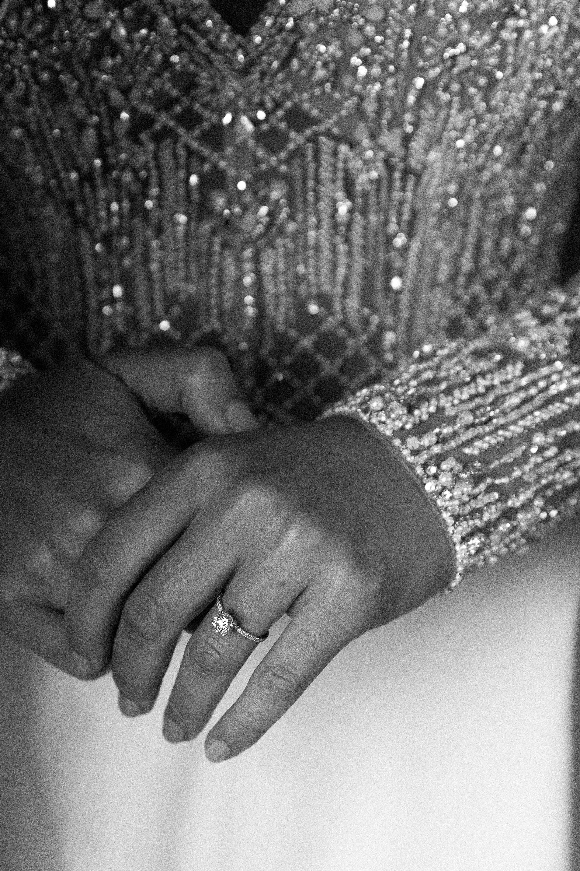 Close-up black and white image of clasped hands with a small diamond ring on the lower hand's ring finger. The background features a textured fabric, likely a sequined dress, creating a sparkling effect. The focus is on the hands and the ring.