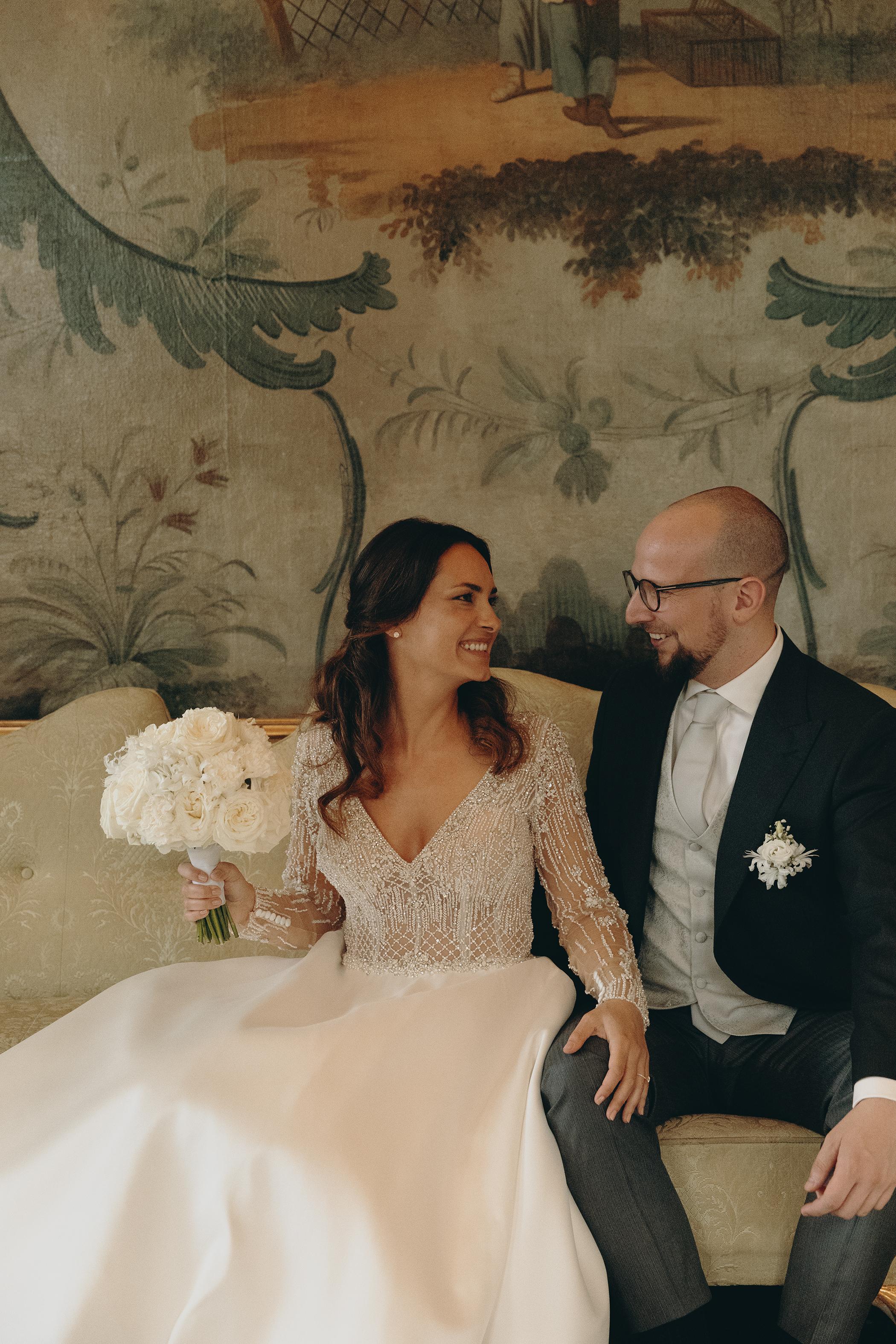 Happy bride and groom seated on a beige couch, smiling and enjoying a tender moment together. The bride wears a cream satin wedding gown with beaded details, holding a bouquet of white roses. The groom is dressed in a dark suit with a light gray waistcoat. The soft, romantic lighting and floral-patterned background add to the intimate wedding atmosphere.