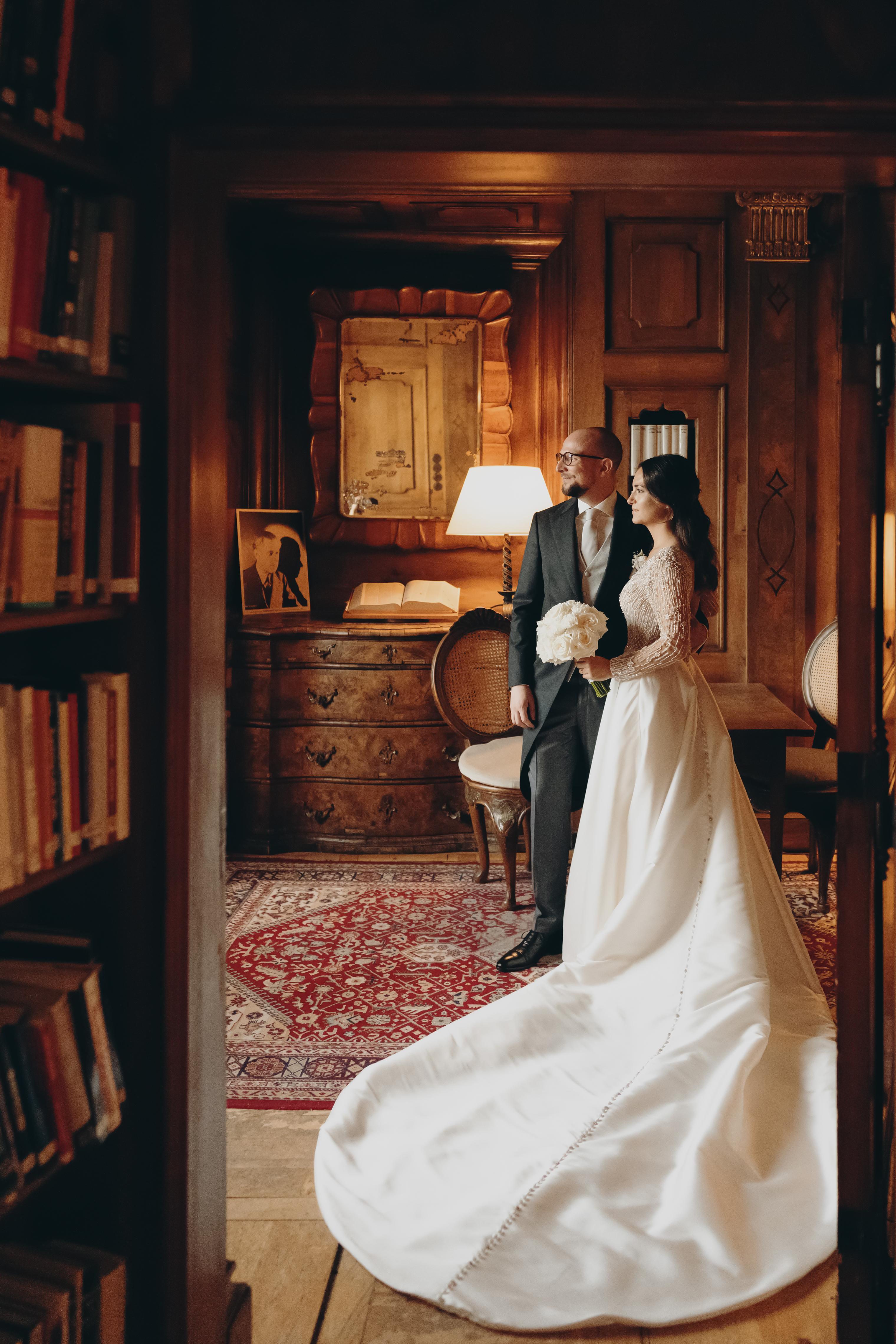 The image shows a bride and groom in an elegant, wood-paneled room decorated with antique furniture and books. The bride is wearing a long white dress, and the groom is wearing a dark suit. They are holding hands and smiling at the camera. The room is warm and inviting, and the atmosphere is romantic and festive. In the background, an antique secretary with books and a mirror can be seen. The red carpet underscores the festive mood.