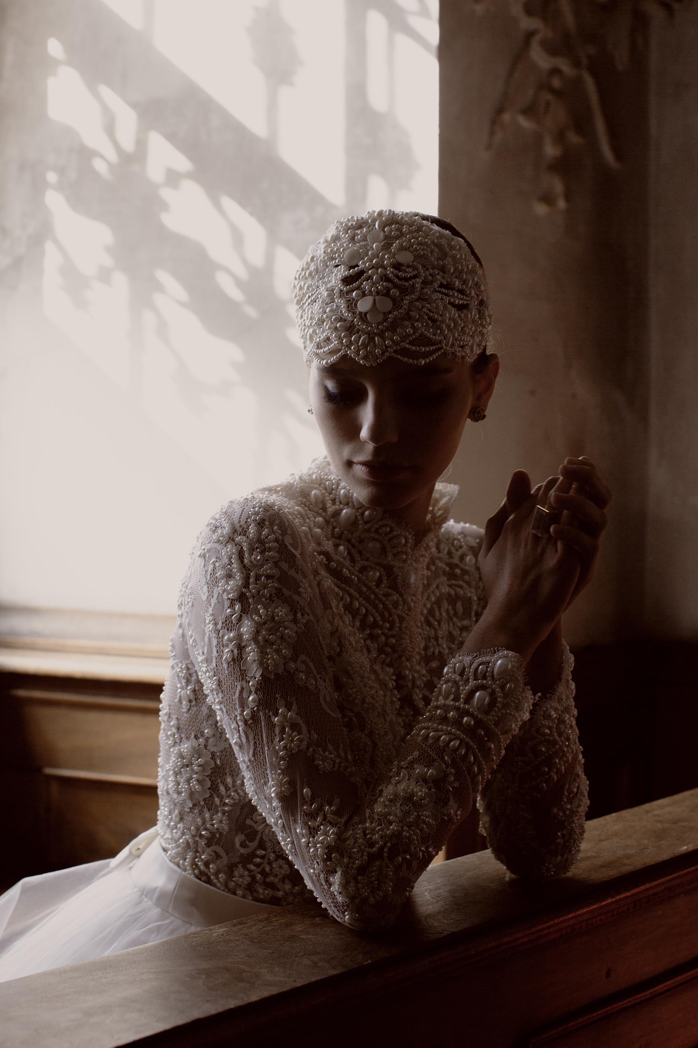 Full shot of a bride in an embroidered, pearl-beaded wedding gown. She gazes downward with eyes closed, wearing an elaborate headpiece adorned with pearls and shells. The gown's bodice and sleeves are richly embroidered with floral and geometric patterns. Her hands are clasped, showing a ring. The soft, warm-toned background features light and shadow effects, focusing attention on the intricate details of the dress and headpiece.