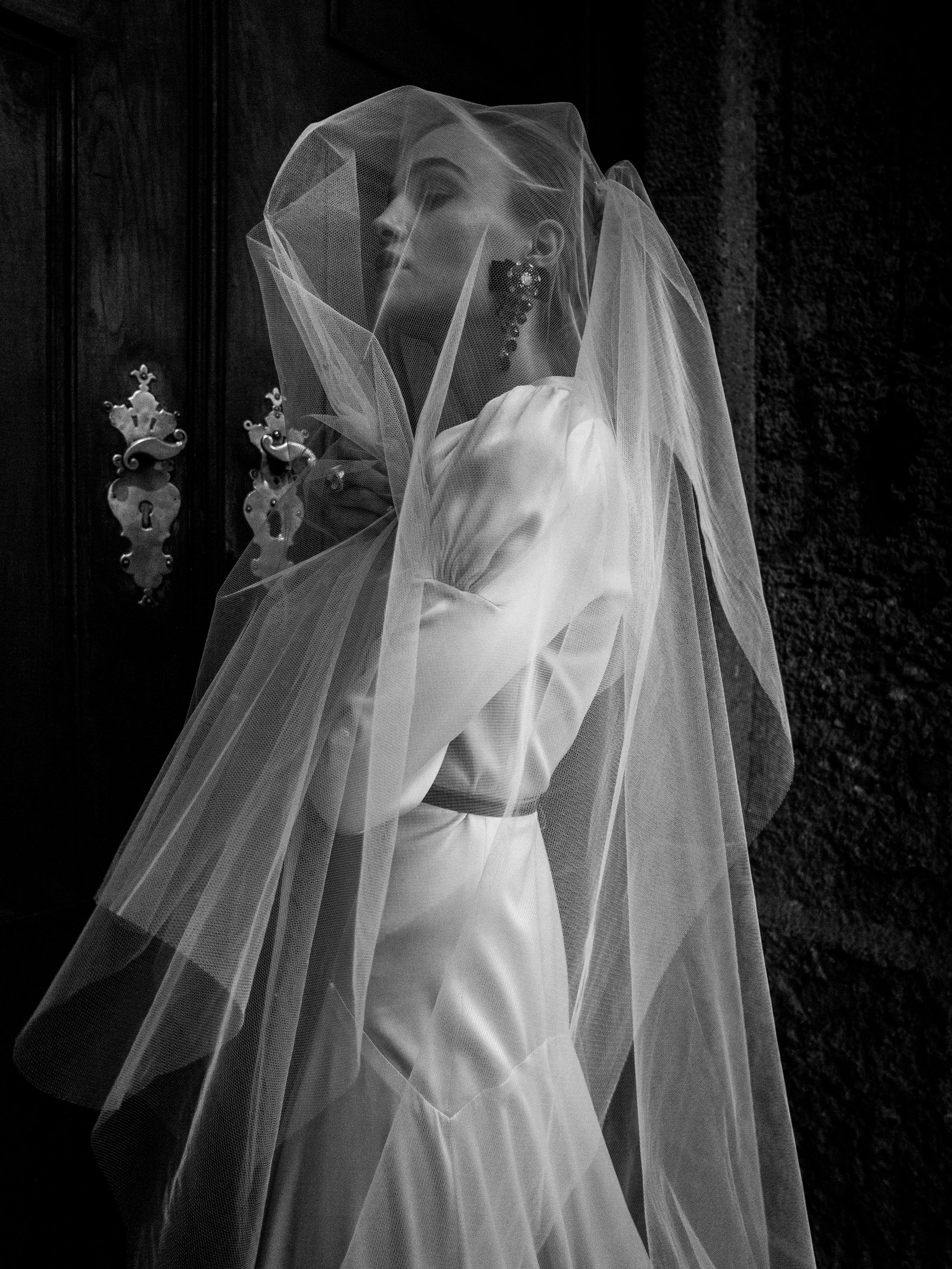 Black and white shot of a bride in a satin wedding dress and veil. The veil drapes around her shoulders, partially obscuring her head. The dress has a flowing, architectural design with a thin sash at the waist. The background is dark and out of focus, with soft light highlighting the bride's form.