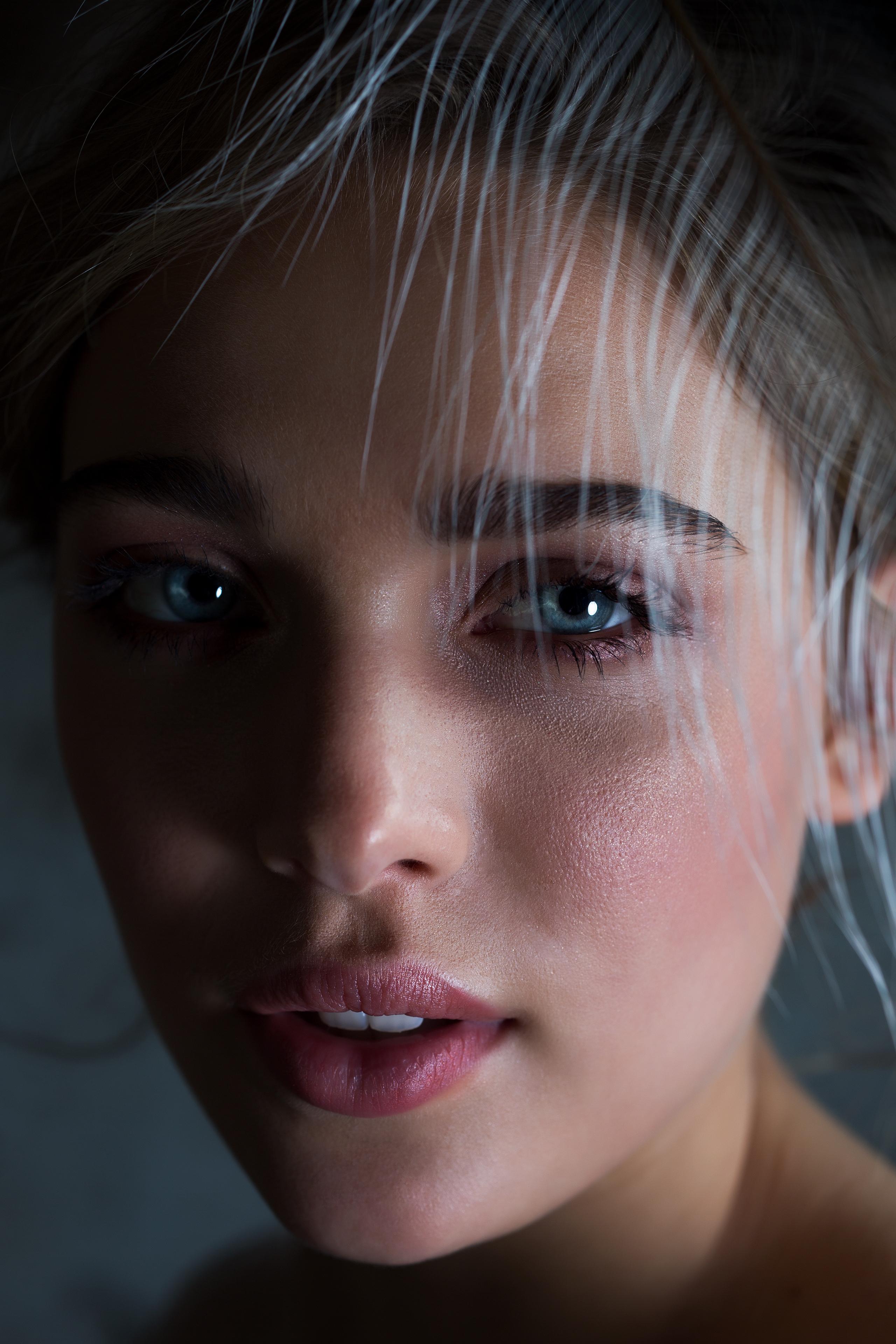 Close-up of a bride's face with light skin and blue eyes. Her expression is neutral with slightly parted lips. She has subtle makeup, including a rosy-pink lipstick and well-defined eyebrows. A light, feathery material drapes across her forehead, adding a soft, ethereal touch. The background is dark, making her features stand out.